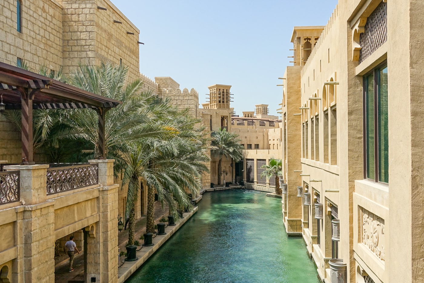 A waterway between traditional Emirati buildings in Dubai, wind
                          towers above the rooftops and date palms along the bank.